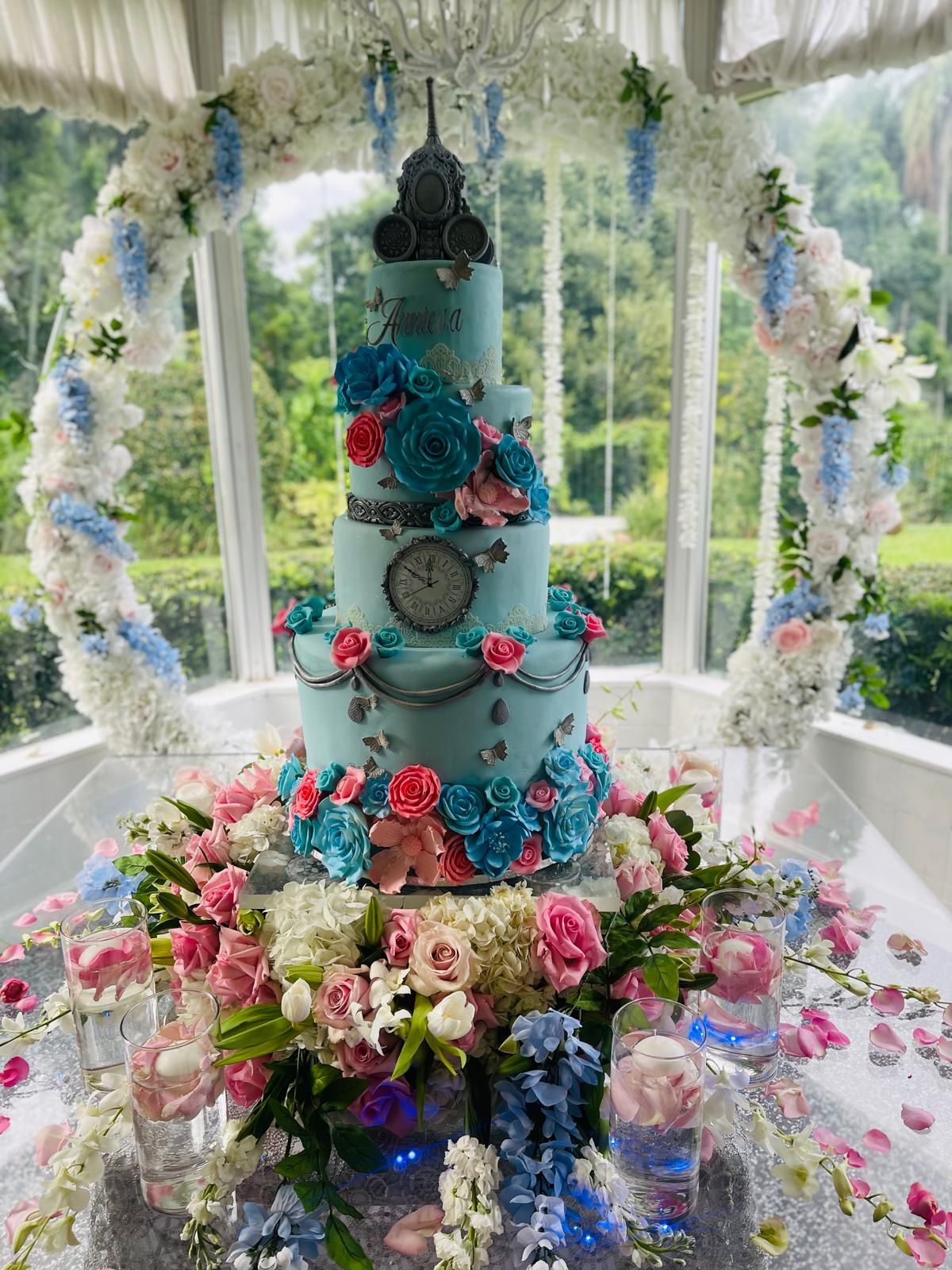 Tall blue wedding cake with flowers and butterfly decorations on a table under a white canopy outdoors