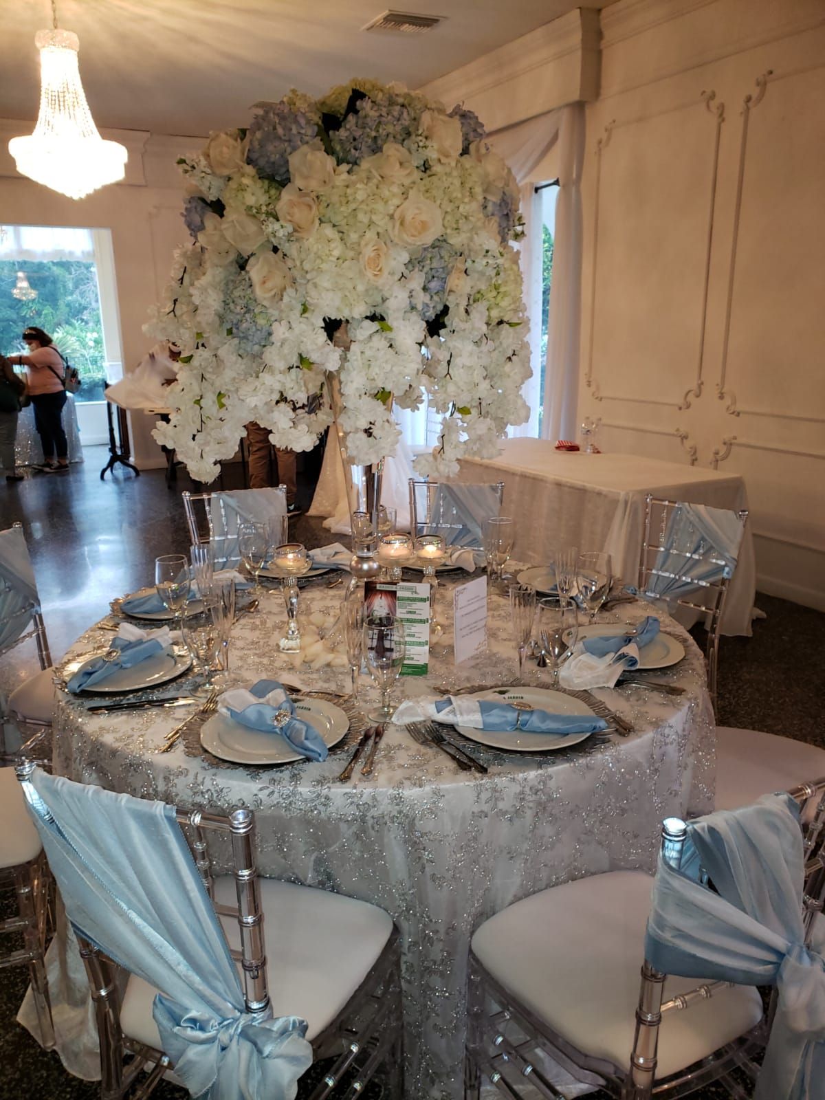 Elegant banquet table with white floral centerpiece, silver decor, and pale blue chair sashes in a formal room