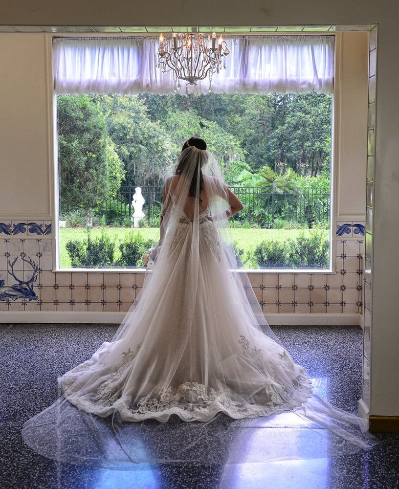 Bride in a long white veil and gown stands by a large window overlooking a green garden.