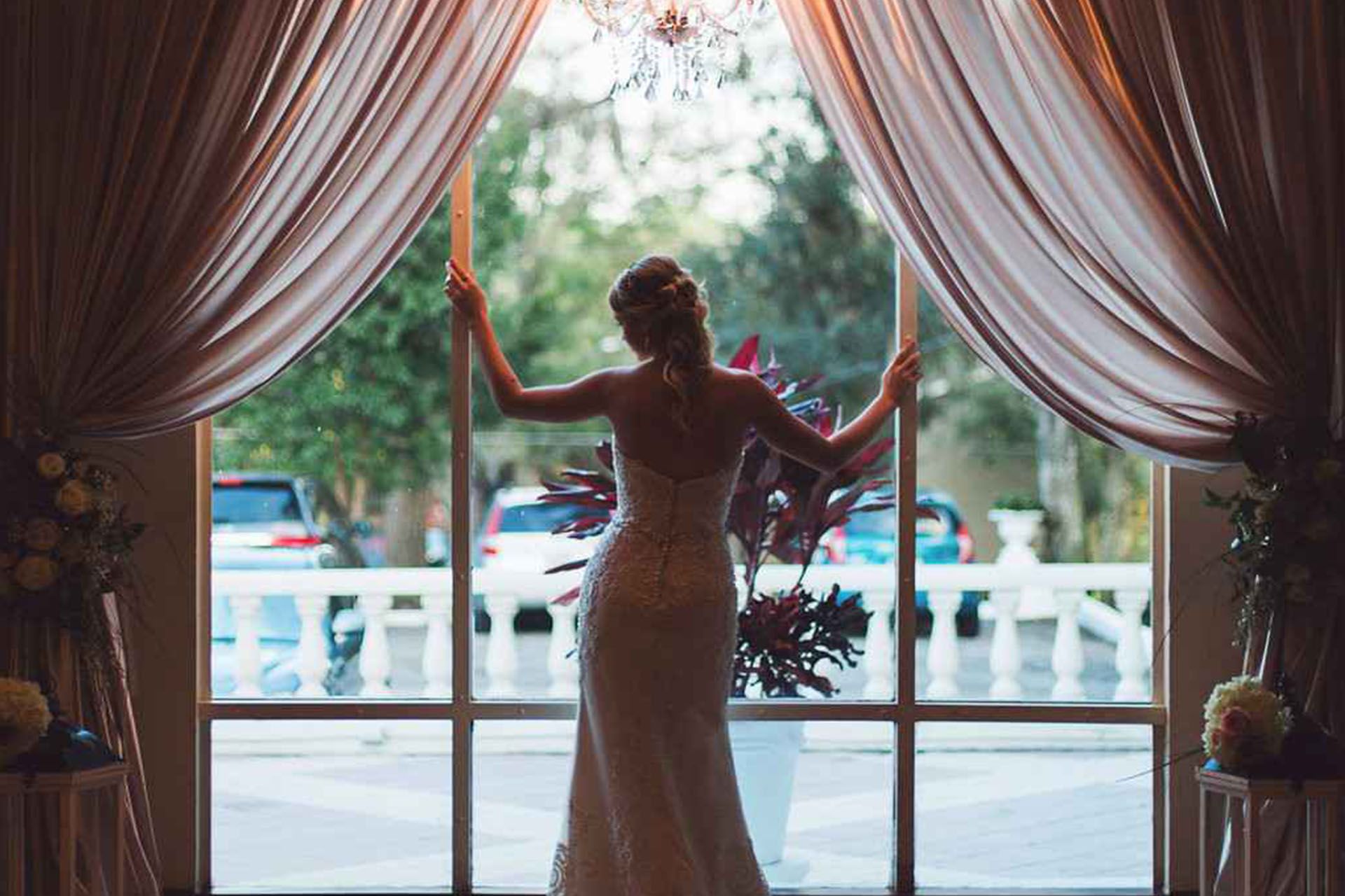 Bride in a white gown stands at a window framed by draped curtains, overlooking a balcony and parked cars.