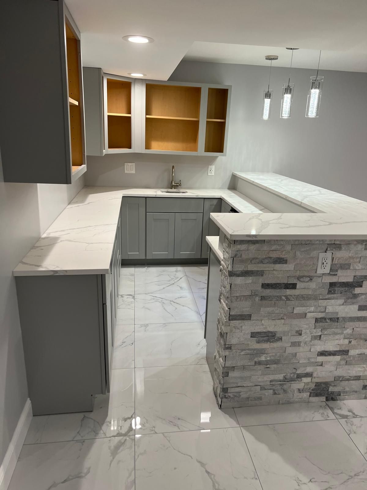 A kitchen with gray cabinets , white counter tops , and a brick wall.