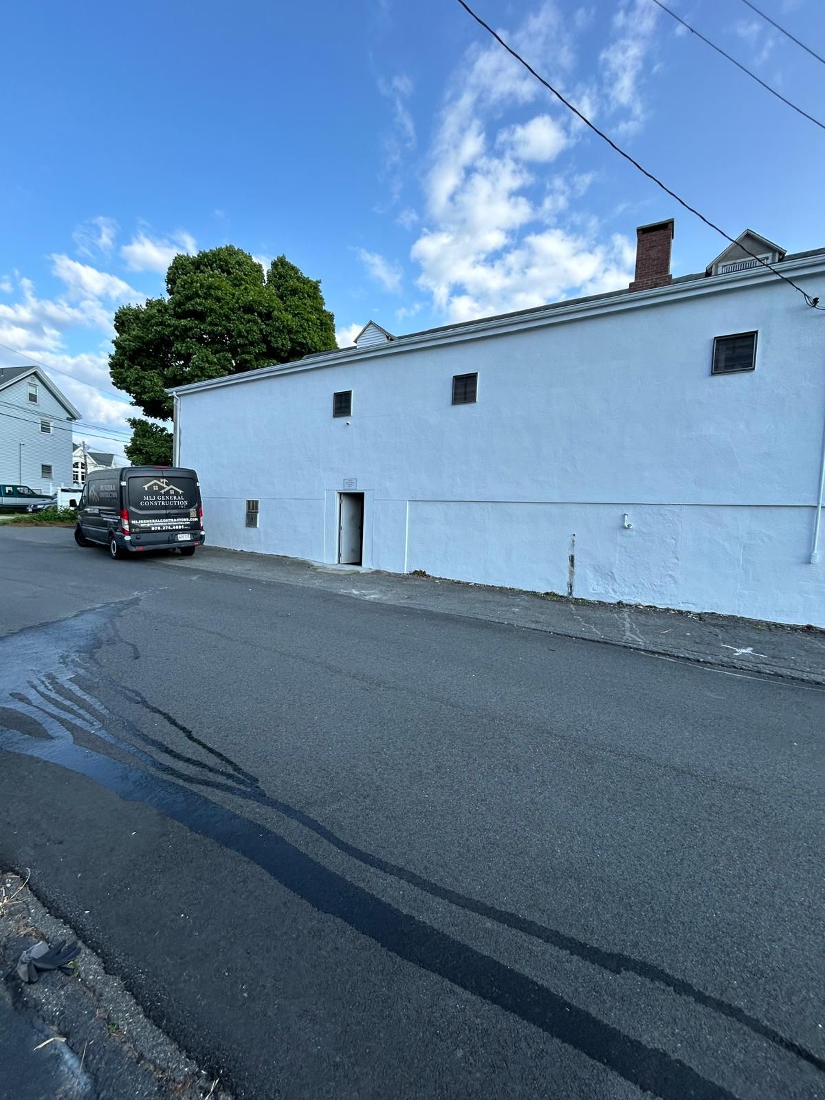 a white building with small windows and a door, next to an asphalt road; van parked on the road