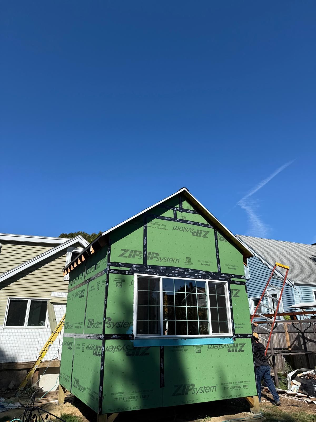 A small, green-wrapped structure with a window under construction against a clear blue sky.