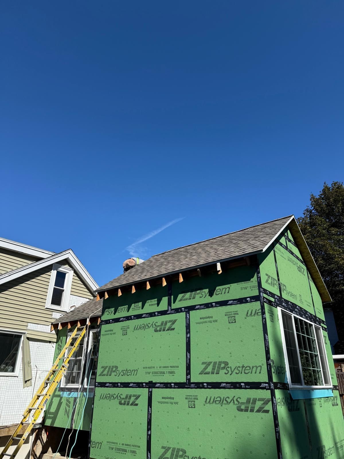 House under construction, green siding, roof partially shingled, ladder leaning against the side, bright blue sky.