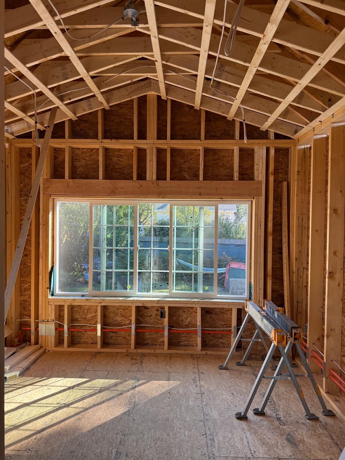 Interior framing of a room with a large window, walls, and rafters. Sunlight streams in.