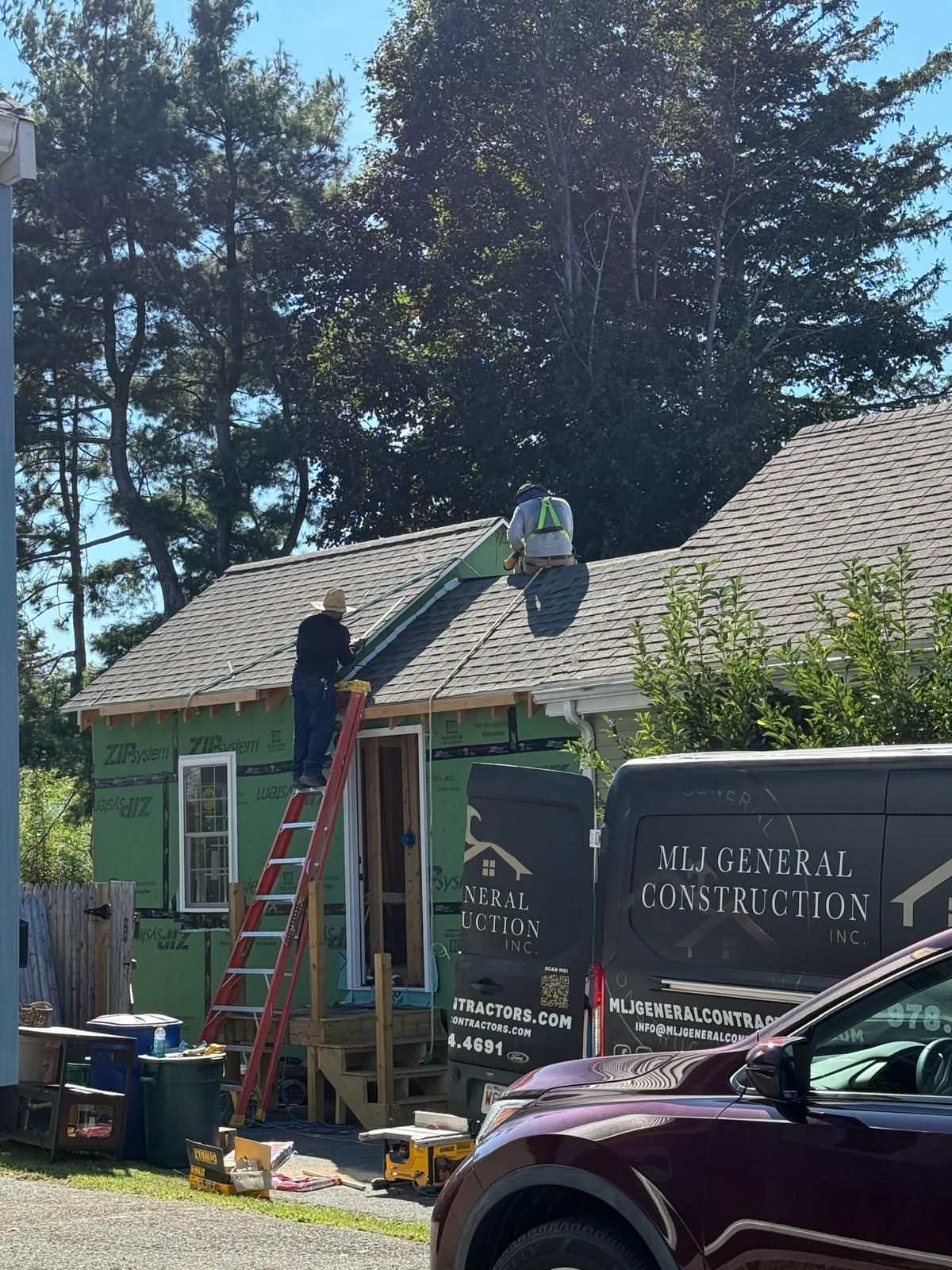 A construction worker on a ladder repairs a house roof with a company truck visible nearby.