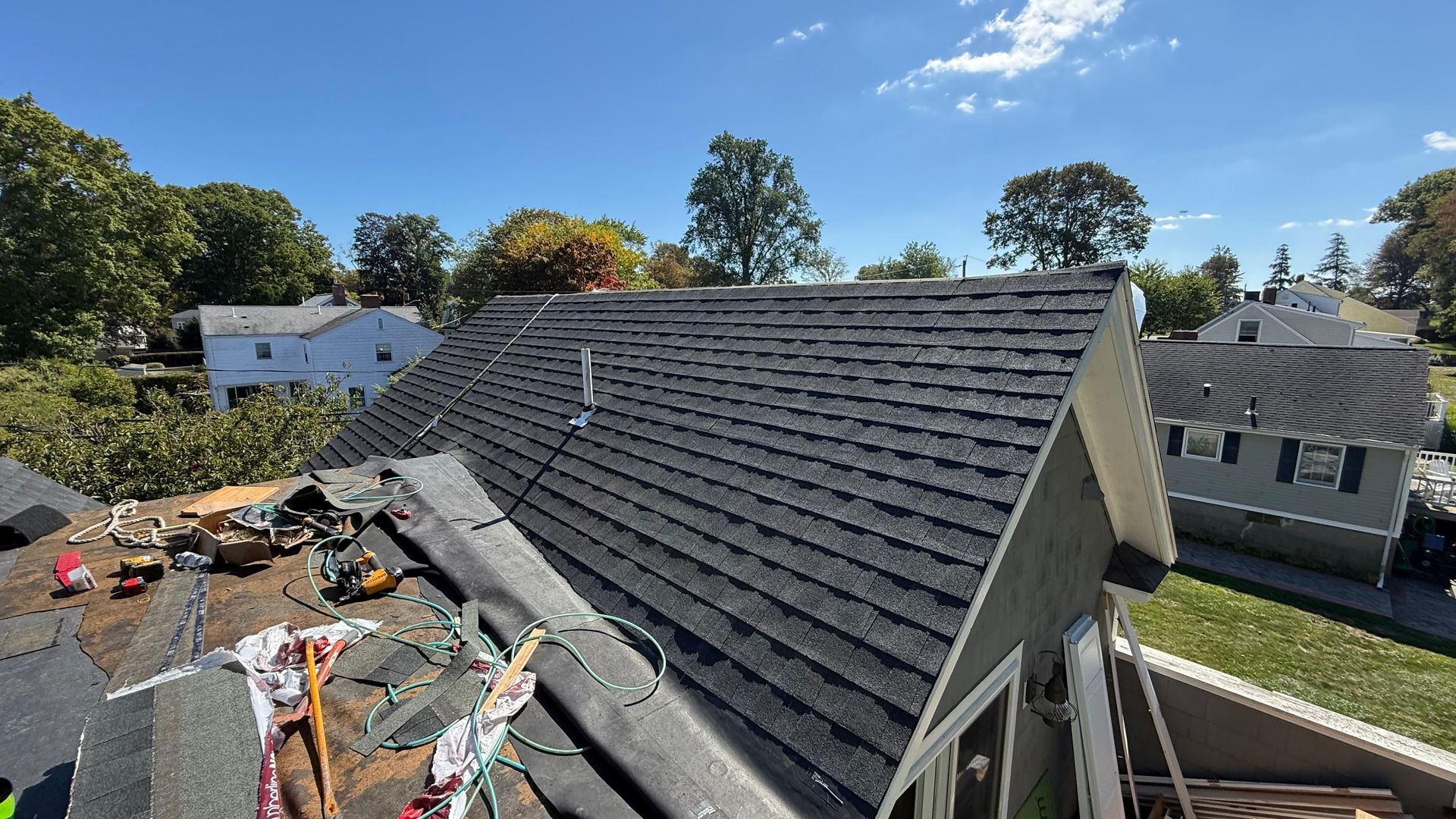 Rooftop with dark shingles, part of it undergoing repair, with trees and houses in the background on a sunny day.