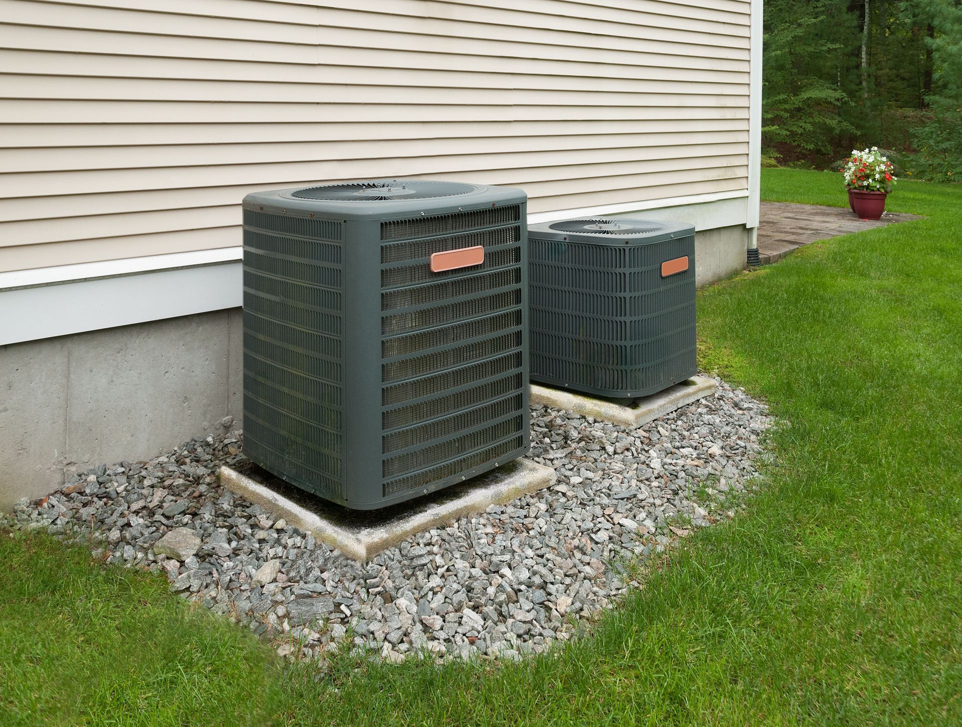 Two gray air conditioning units next to a house on gravel.