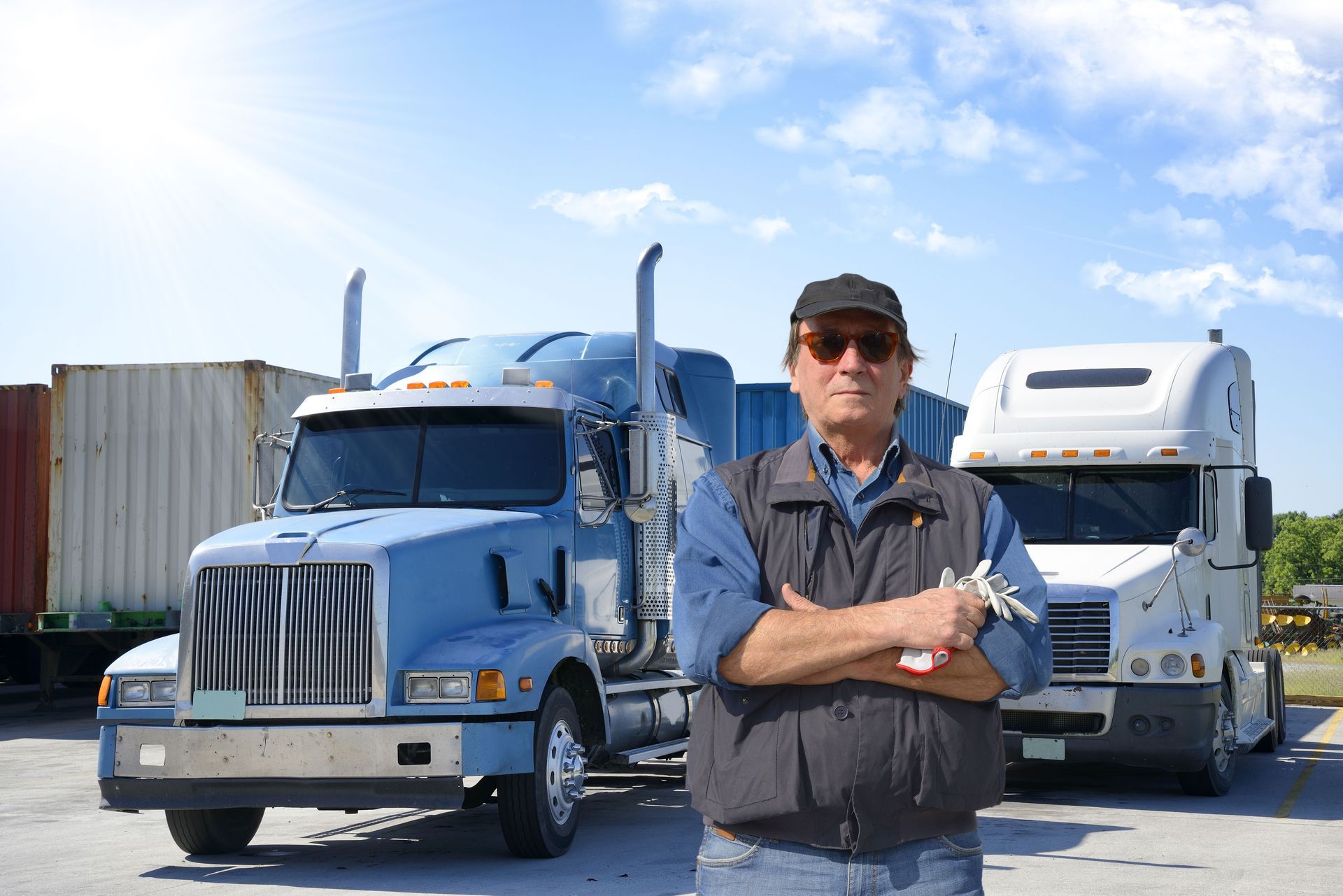 Man in front of two semi-trucks, arms crossed. Sunny outdoor setting.