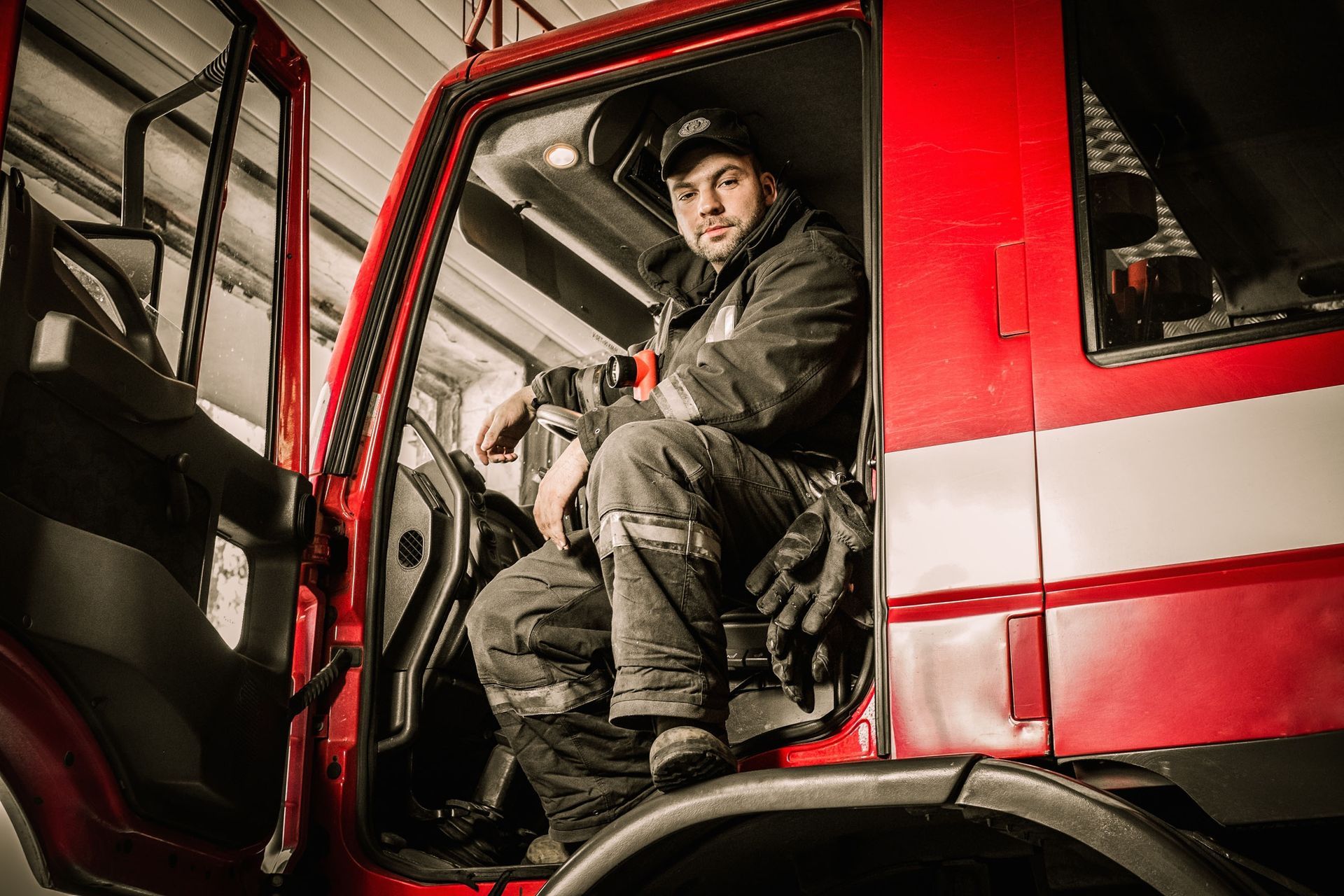 Firefighter in dark work attire sits in the cab of a red fire truck, looking at the camera.