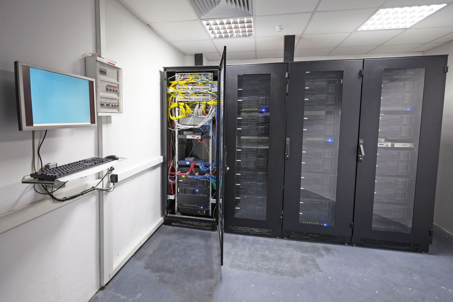 Server room with black racks, open network cabinet, and a wall-mounted monitor on the left.