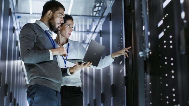 Two technicians in a server room inspecting cables and holding a laptop and clipboard.