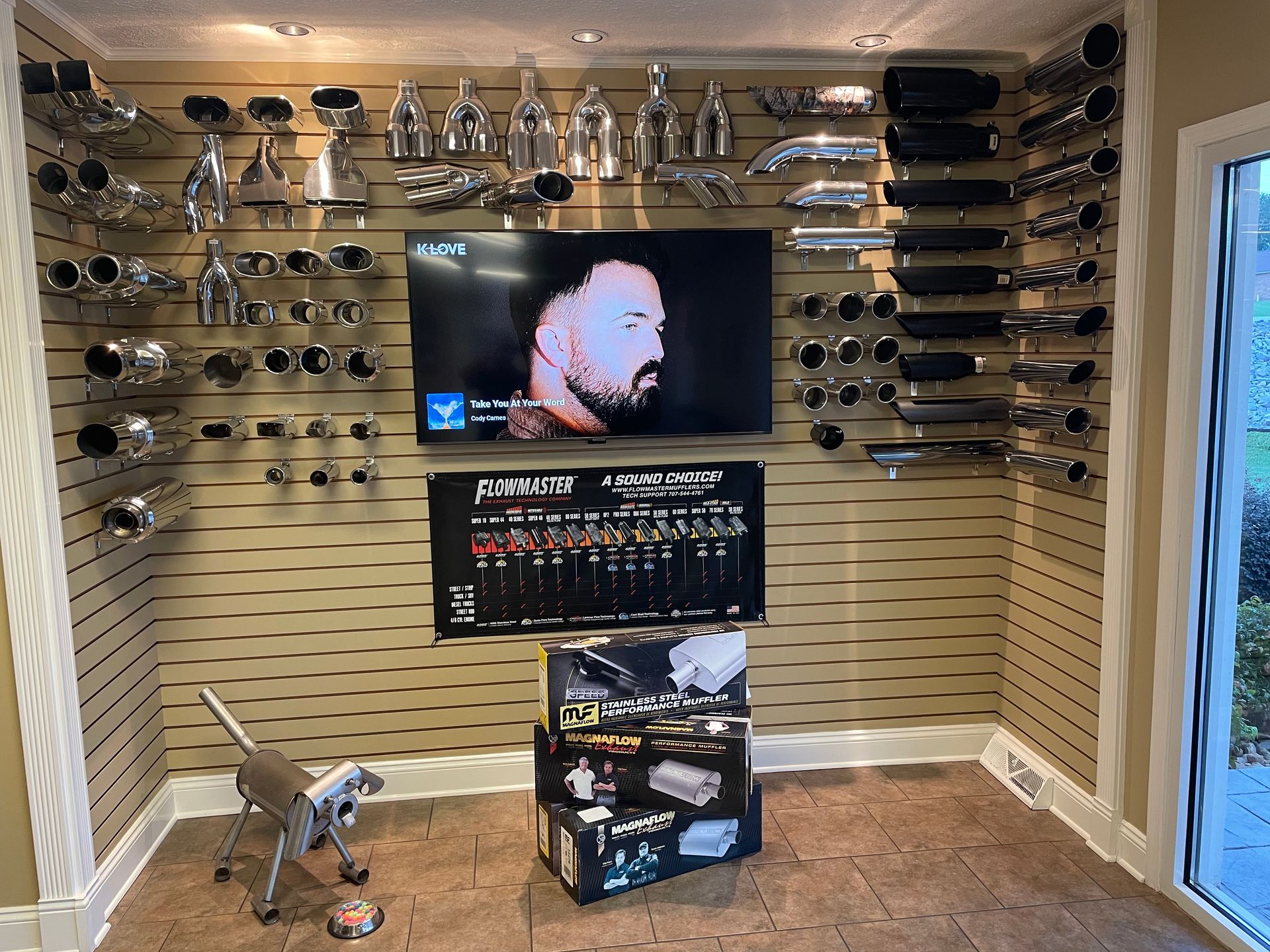 Wall display of exhaust pipes and mufflers; a TV screen shows a person.