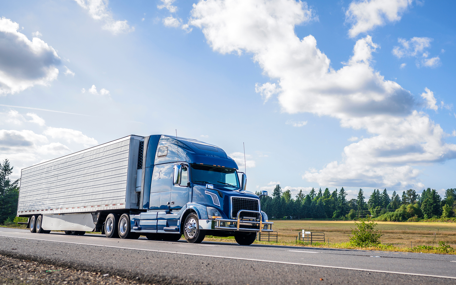 A blue semi truck is driving down a highway next to a field.