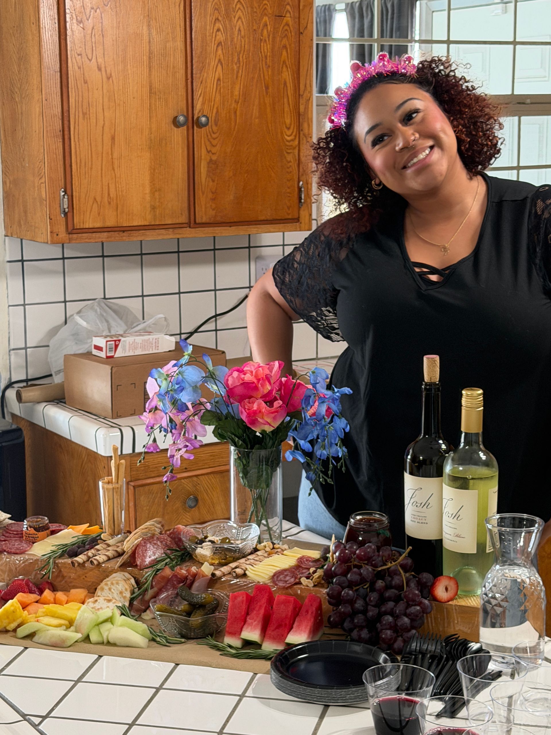 Woman smiling beside a buffet table with fruit, flowers, and wine bottles in a kitchen.