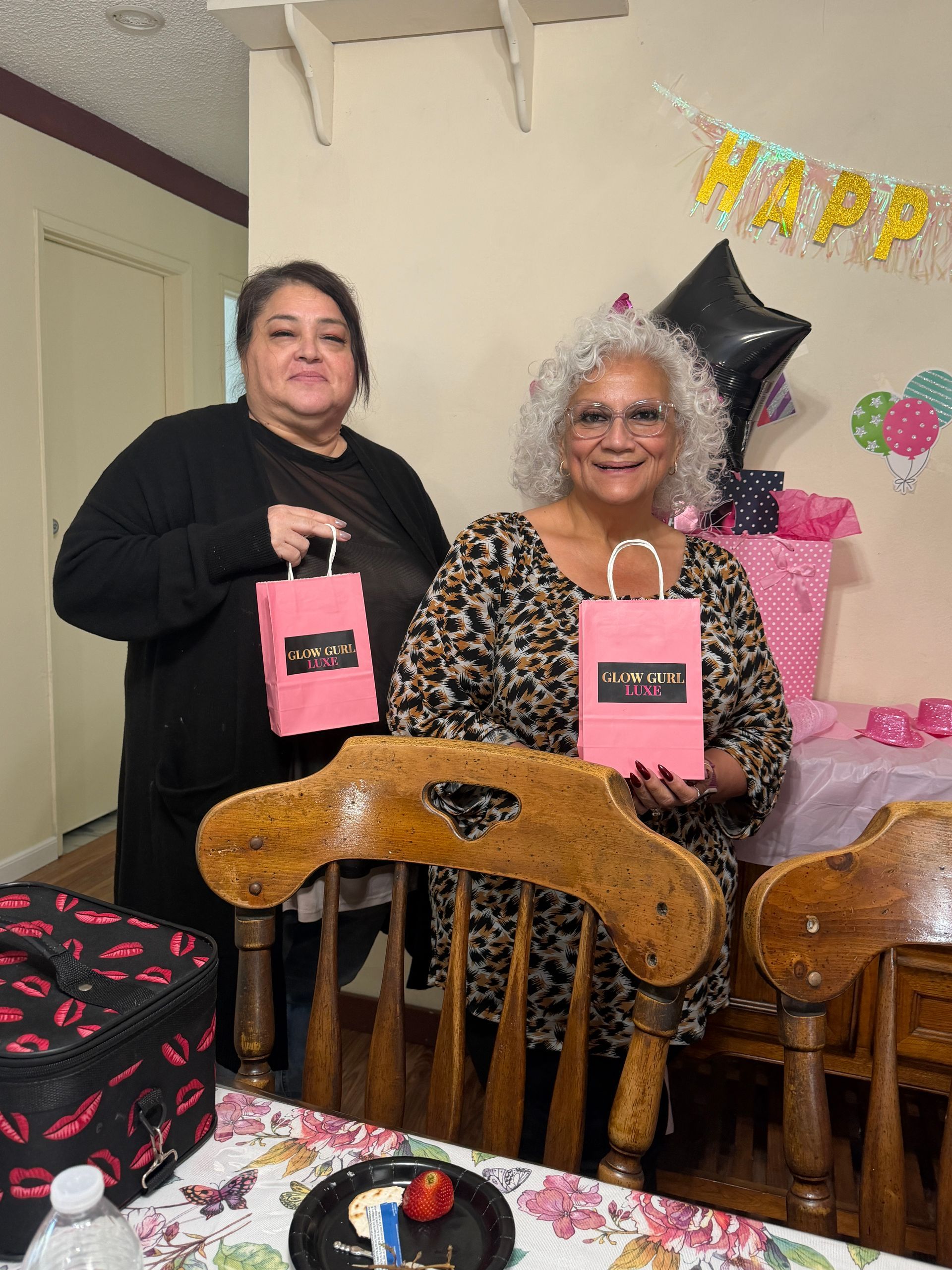 Two women holding pink gift bags at a decorated birthday party table with balloons and a “HAPPY” banner.
