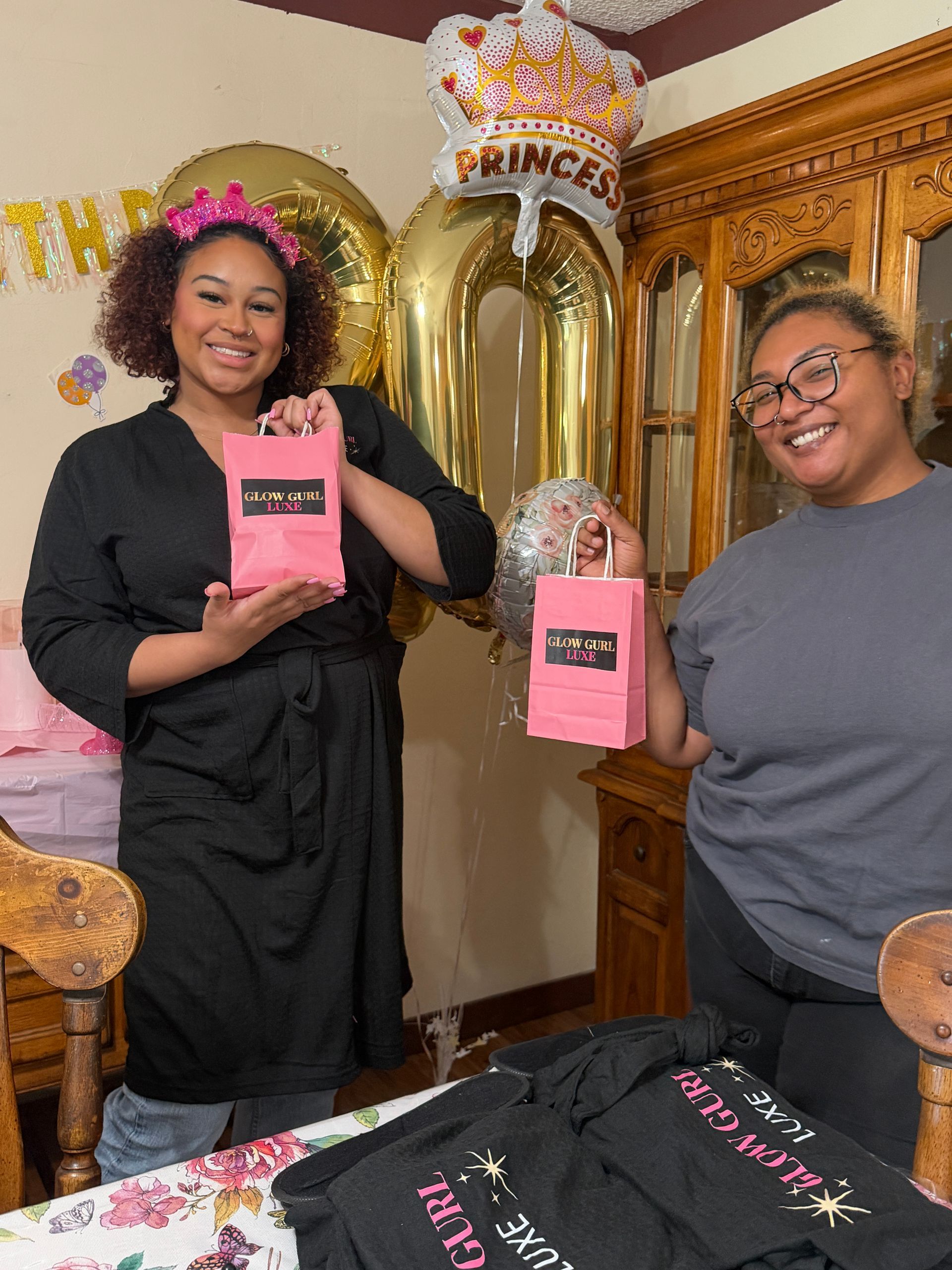 Two people holding pink party favor boxes beside gold balloons and a decorated cake table