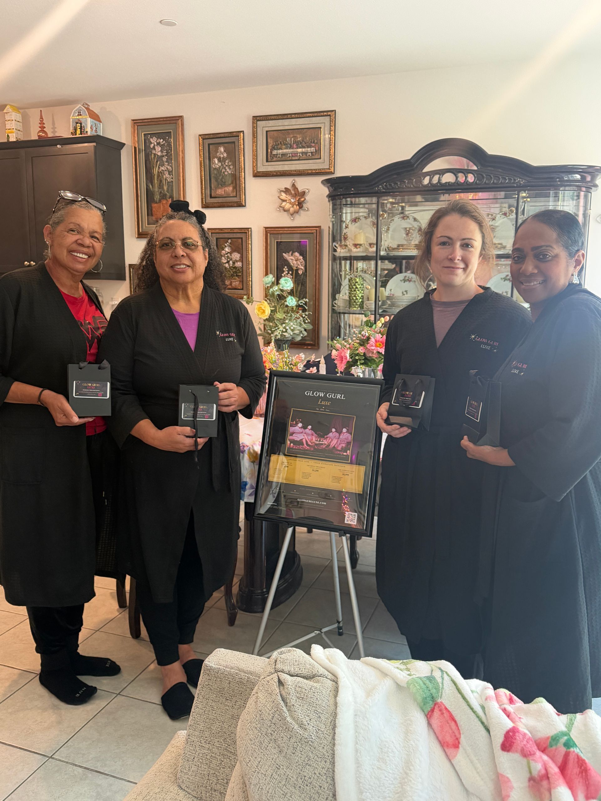 Four women in black outfits stand indoors beside a framed floral display and easel in a decorated room