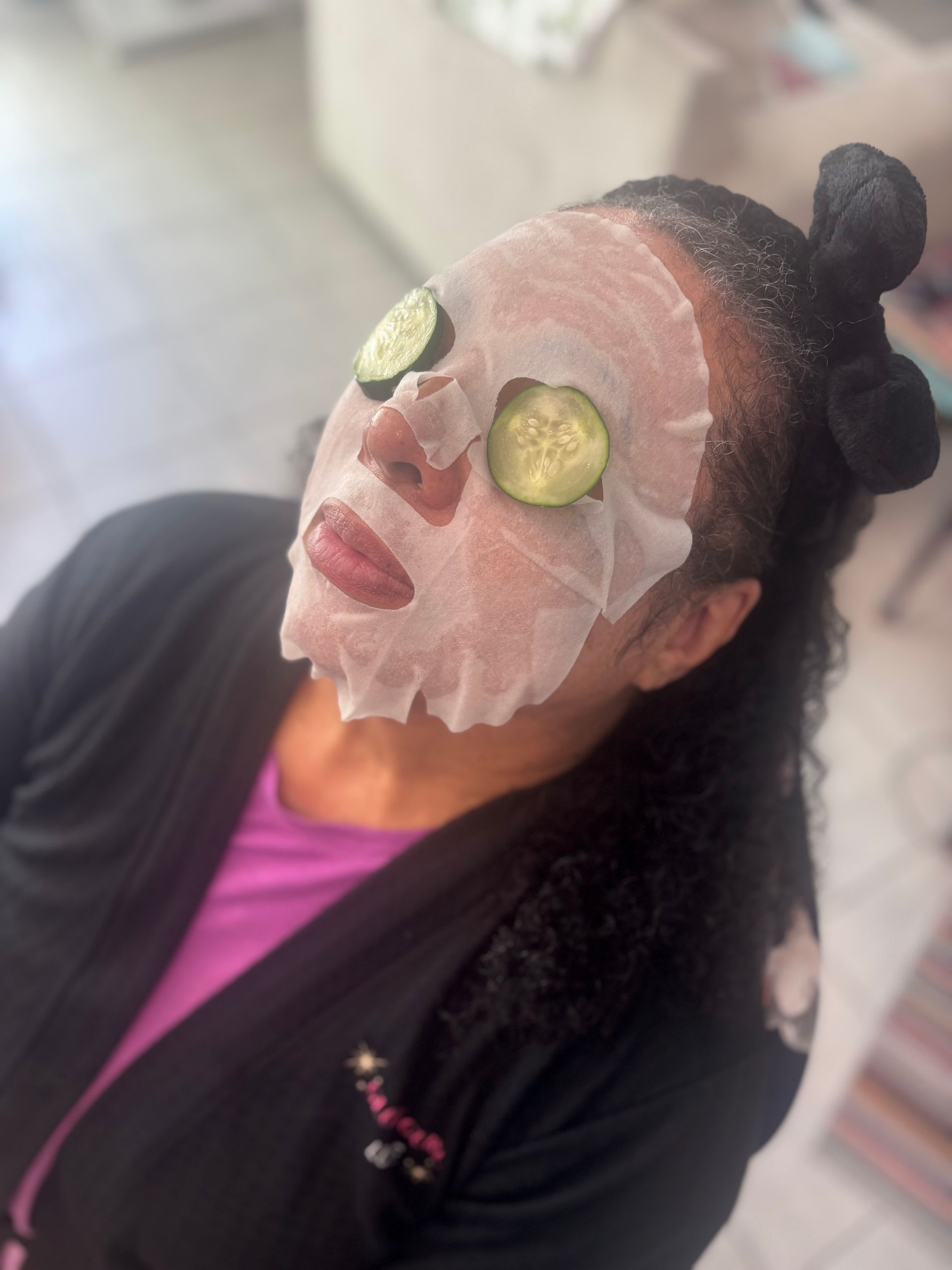 Person with cucumber slices over eyes and a white facial mask, wearing a black jacket and purple shirt.