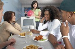 A group of people are sitting at a table eating food while a woman holds a bowling ball.
