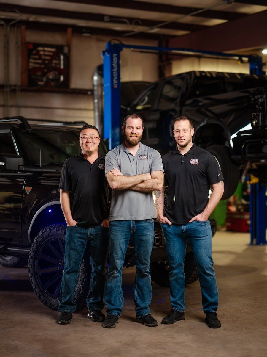Three mechanics posing in a workshop beside a lifted off-road truck