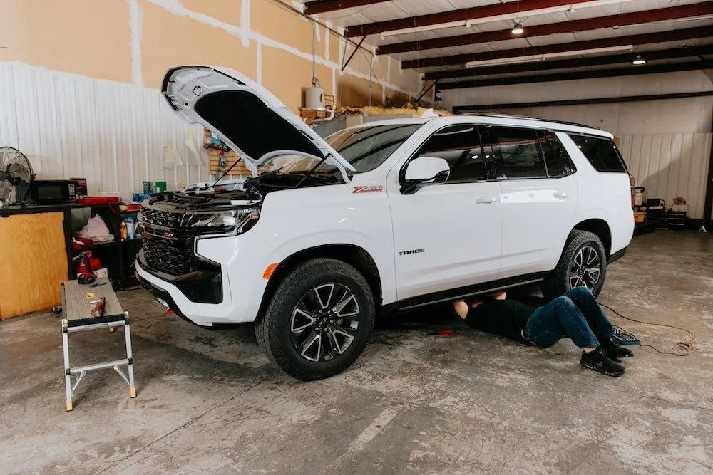 White SUV with hood open in a garage, mechanic working underneath on the passenger side.
