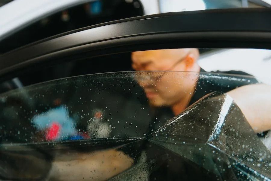Person seen through a rain-speckled car window, with a blurred interior and dark frame.