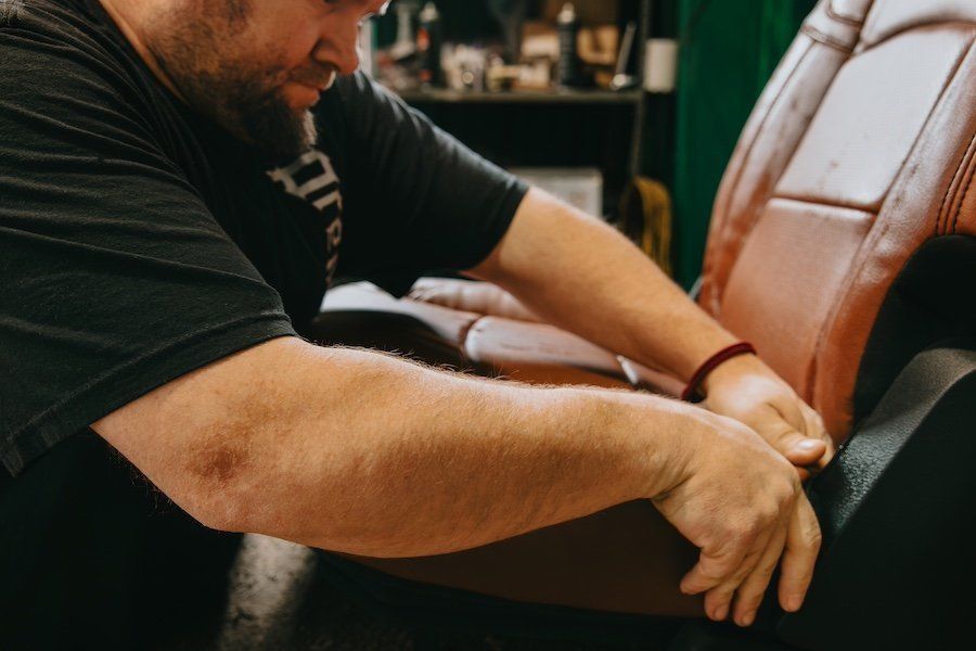 Man in black shirt working with hands near a brown leather chair in a dim workshop