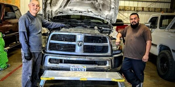 Two men are standing next to a truck with the hood up in a garage.