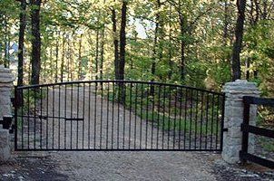 A wrought iron gate is open to a gravel road in the woods.