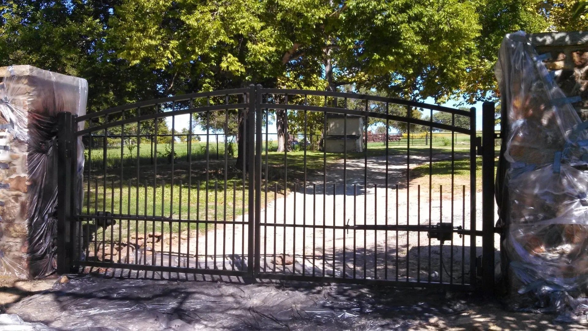 A wrought iron gate is open to a driveway with trees in the background.