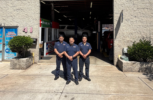 Three people in work uniforms stand in front of National Motorworks Auto Repair shop under a clear blue sky.