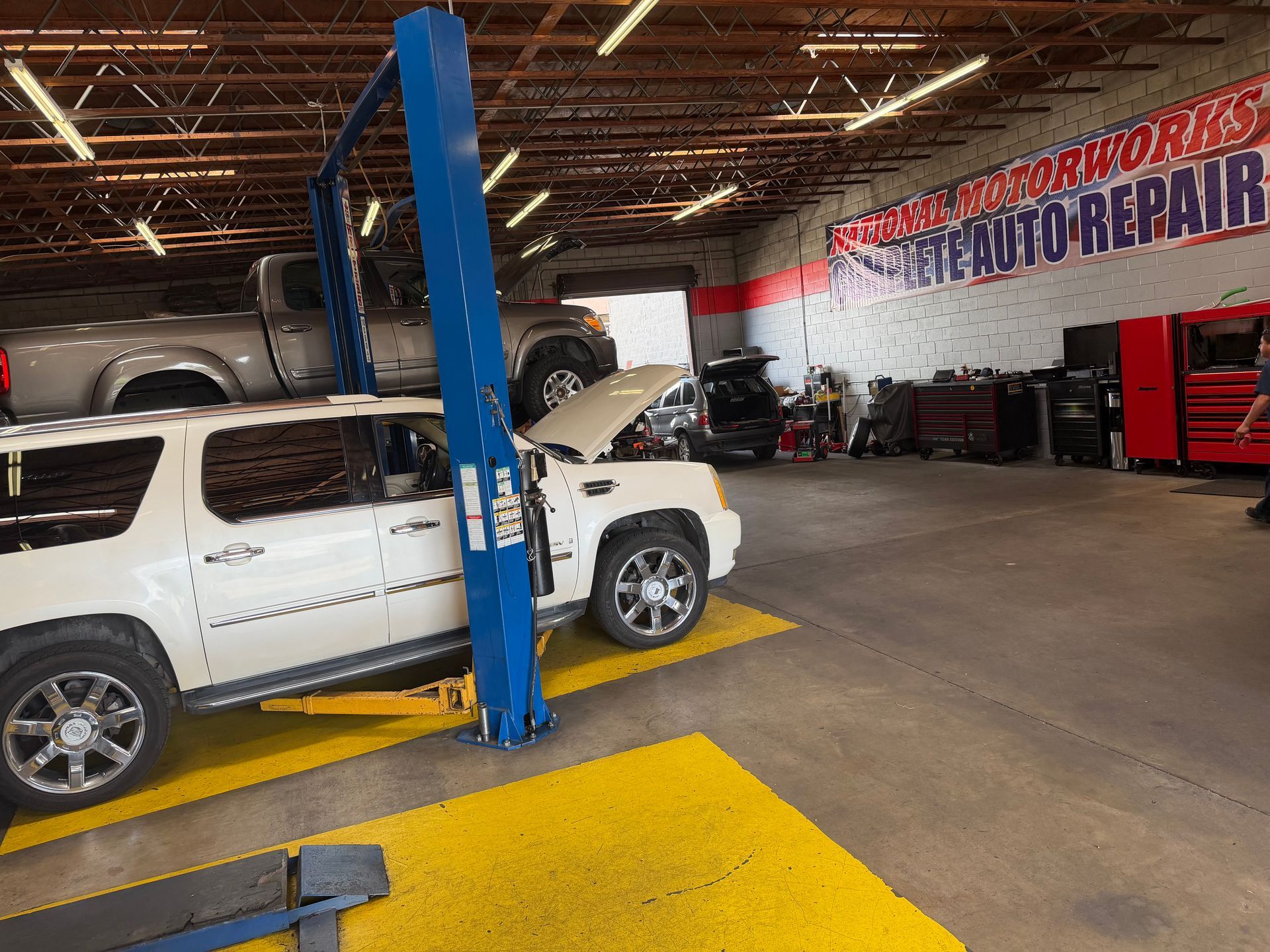 A white SUV lifted on a garage hoist inside an auto repair shop with a large wall sign and red tool cabinets.