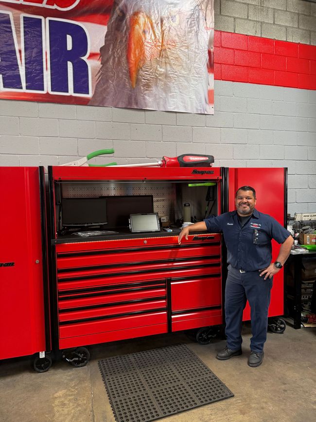 A smiling mechanic in a uniform standing beside a large, red professional tool chest in a garage.