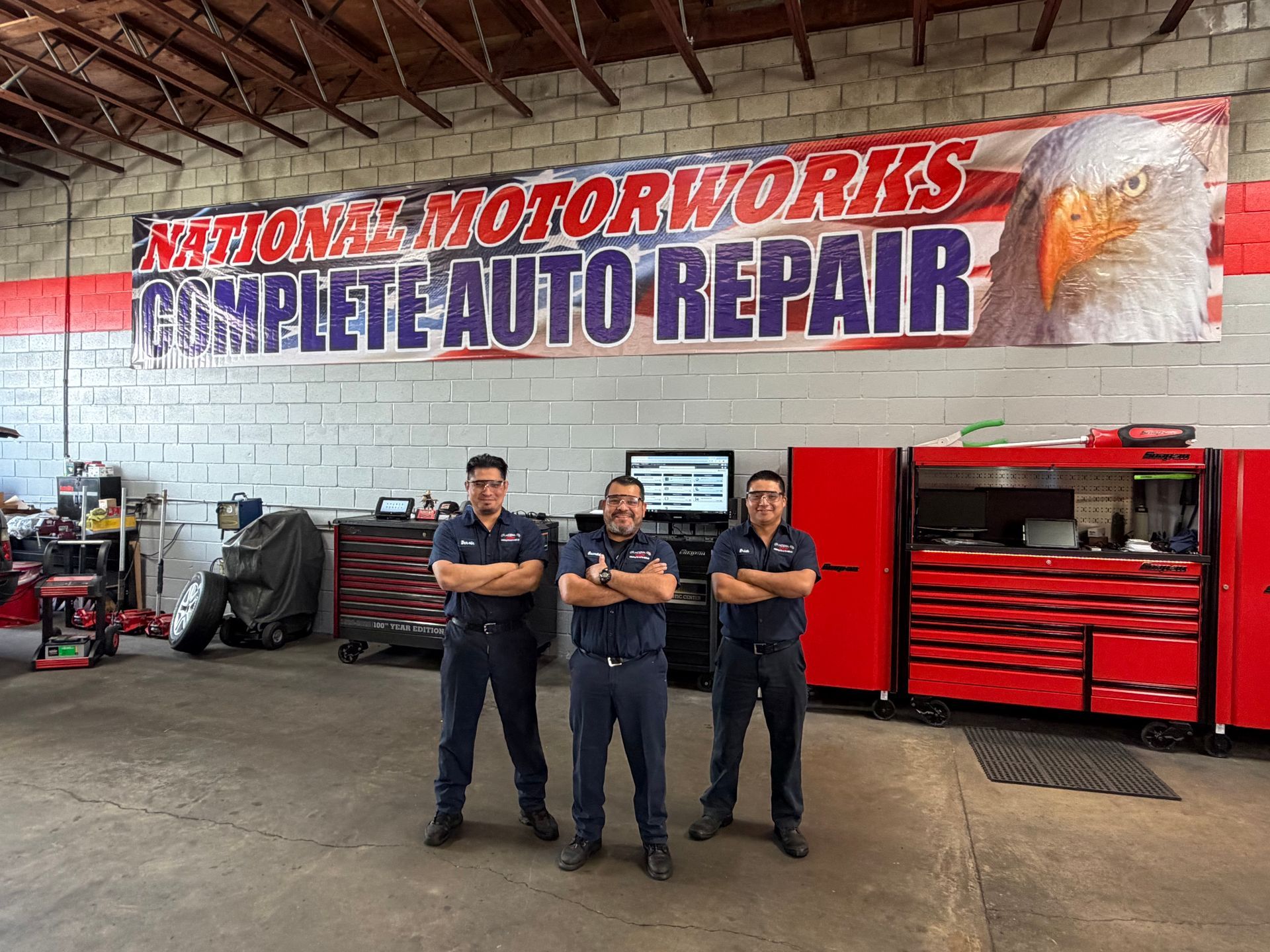 Three mechanics stand with arms crossed in an auto repair shop in front of a banner for National Motorworks.