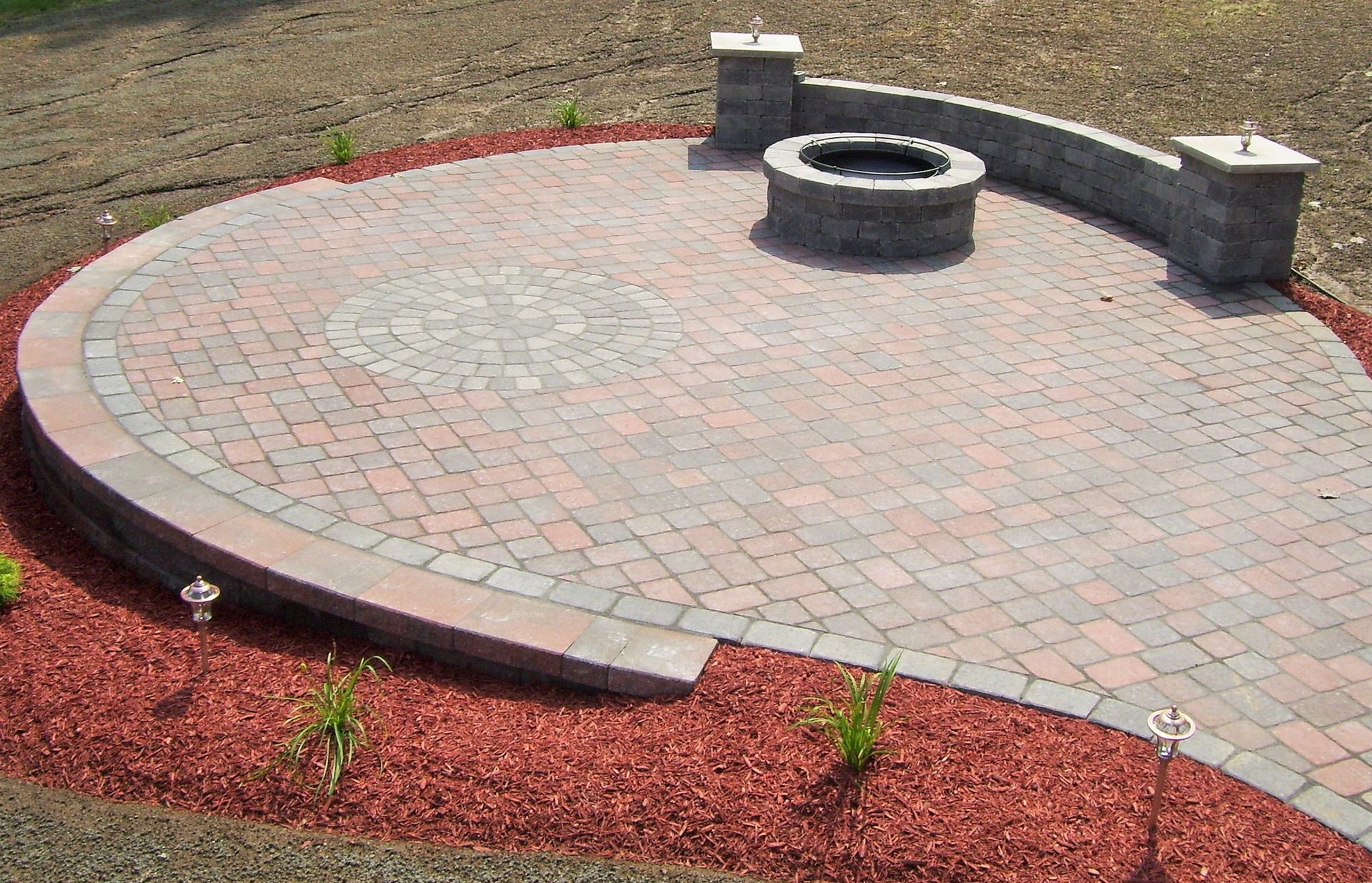 A circular stone patio with a fire pit, curved seating wall, and pillars, surrounded by red mulch.