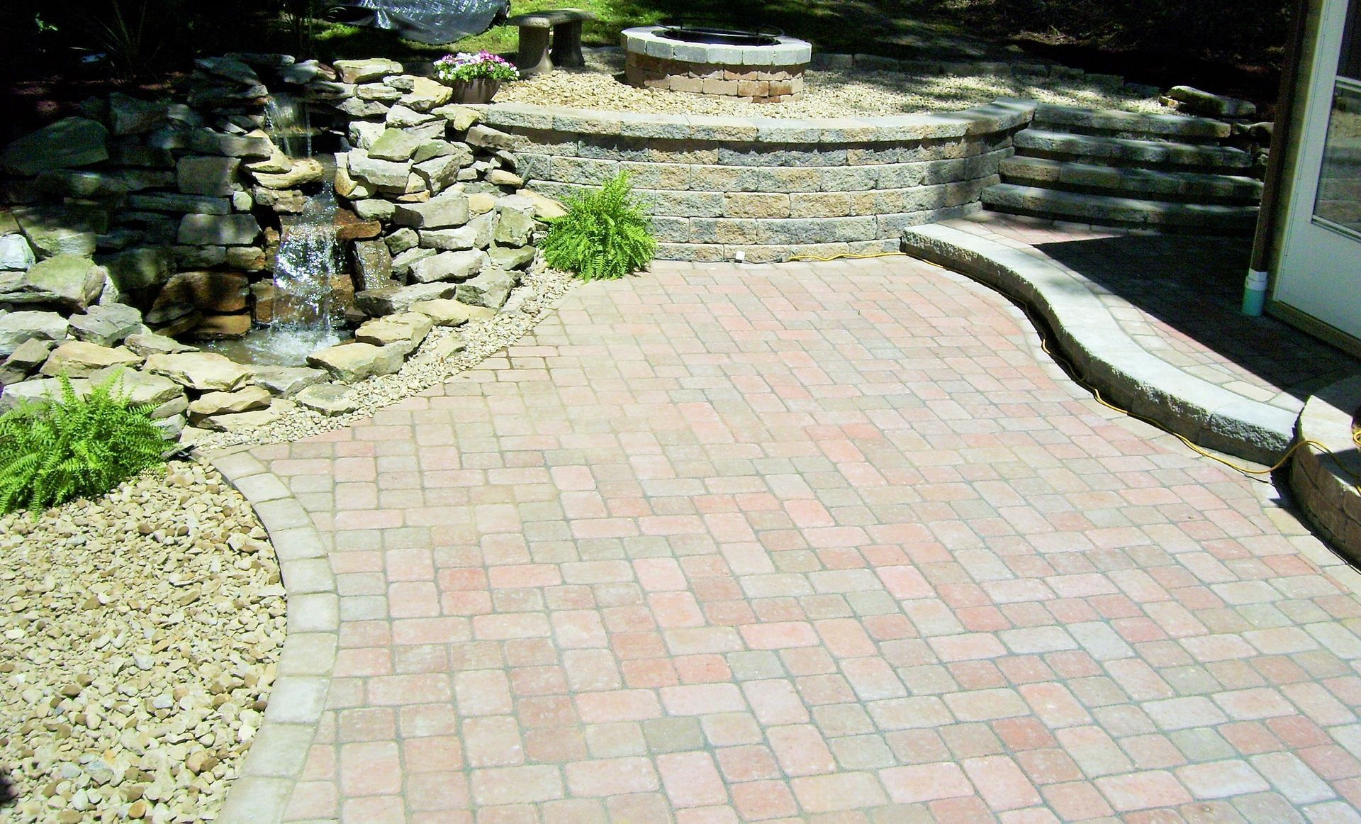 A stone paver patio with a waterfall, a stone retaining wall, and a circular fire pit area in a garden setting.