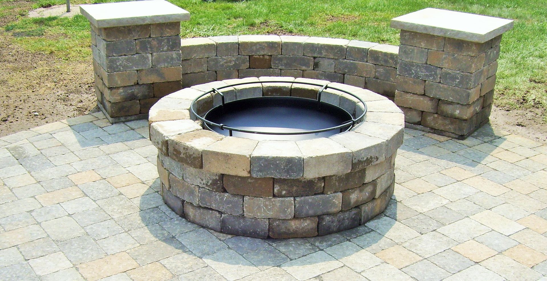 A circular stone fire pit sits on a paved patio with a curved stone wall and two pillars in the background.