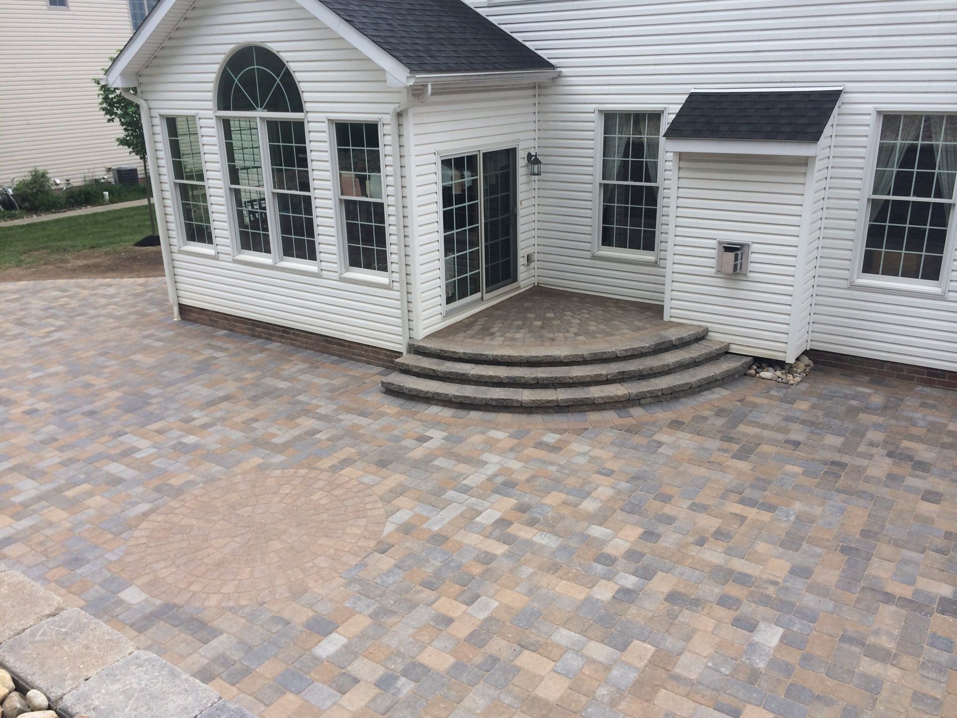 A beige and gray paver patio with a circular design in front of a white house with stone stairs leading to a door.