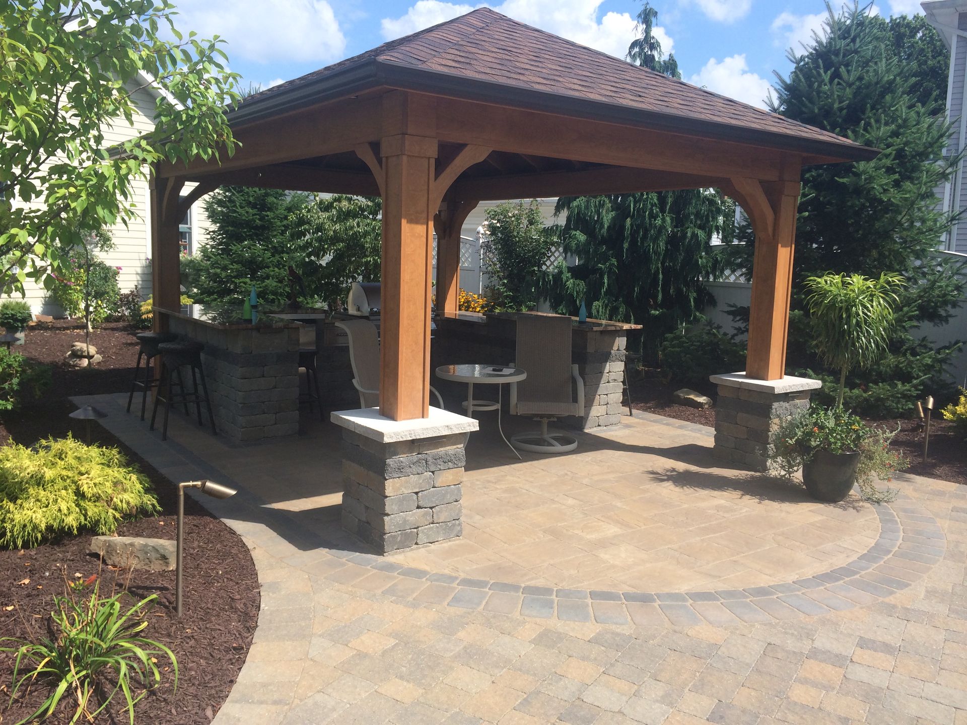 A wooden pavilion with a dark roof over a stone patio featuring a built-in outdoor kitchen, bar stools, and a small table.