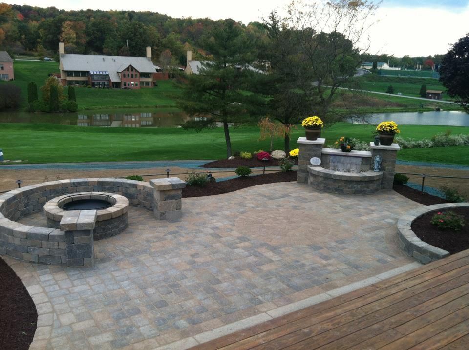 A backyard stone patio featuring a circular fire pit, a stone bench, and landscaping, overlooking a pond and green hills.