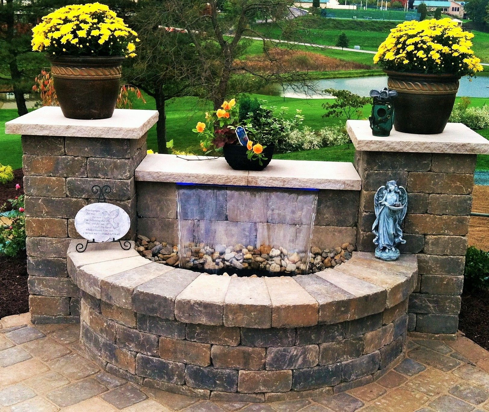 A brick-built water feature with two planters of yellow chrysanthemums and a small angel statue in an outdoor garden.