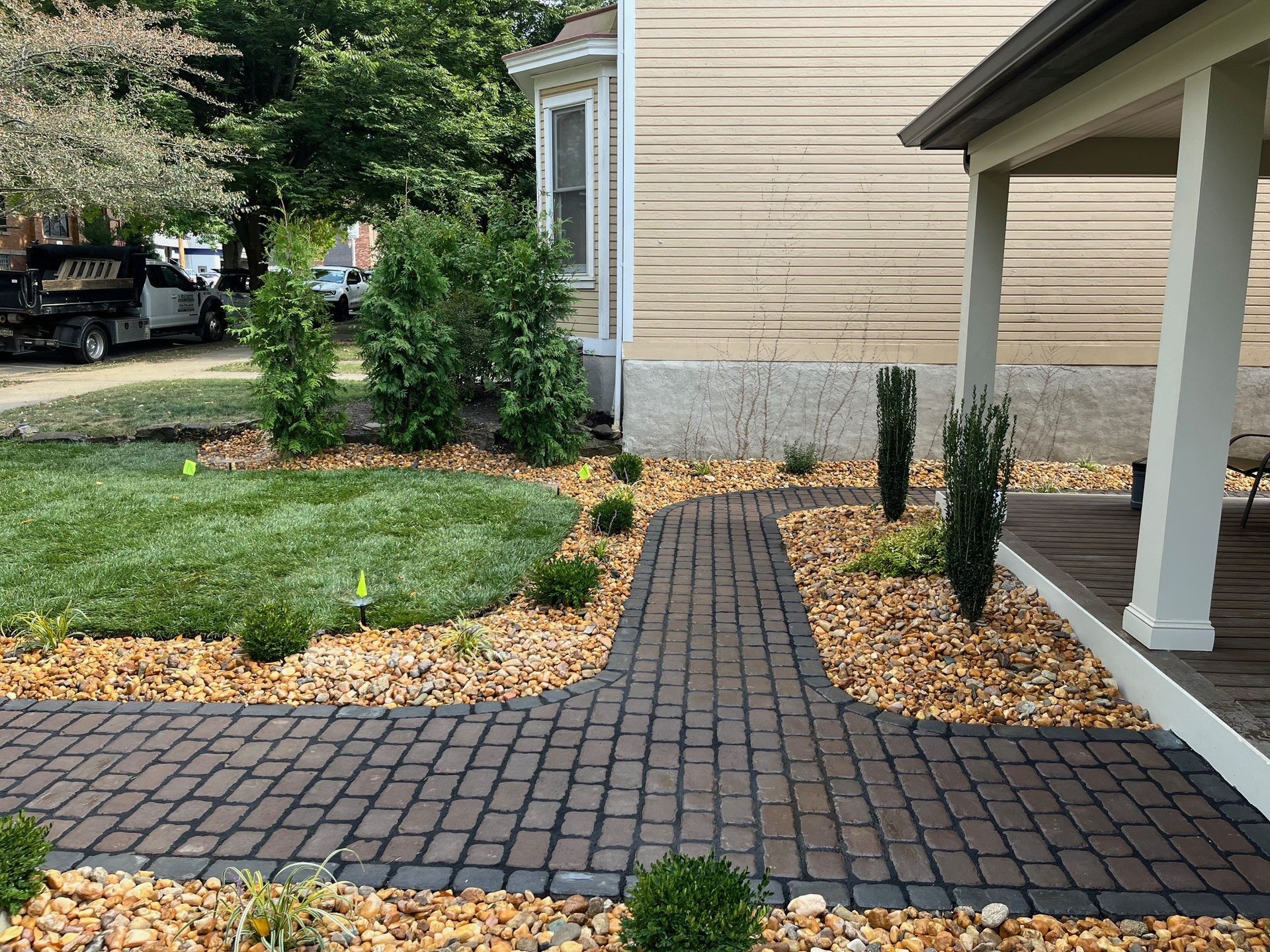 A dark brick pathway winds through a front yard lined with tan gravel, small shrubs, and a white covered porch.