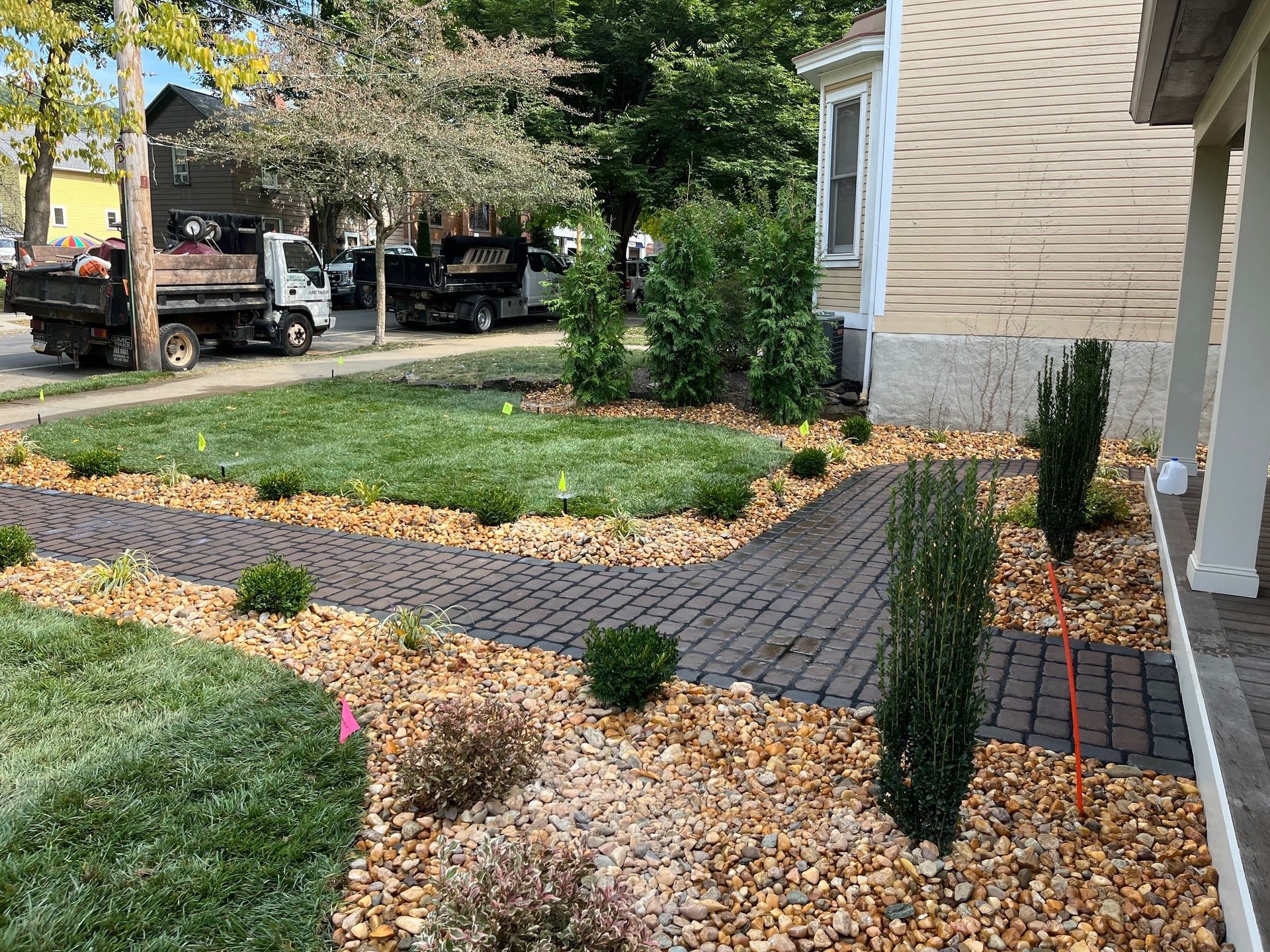 A brick walkway curves through a front yard with mulch beds, small shrubs, green grass, and trucks in the background.