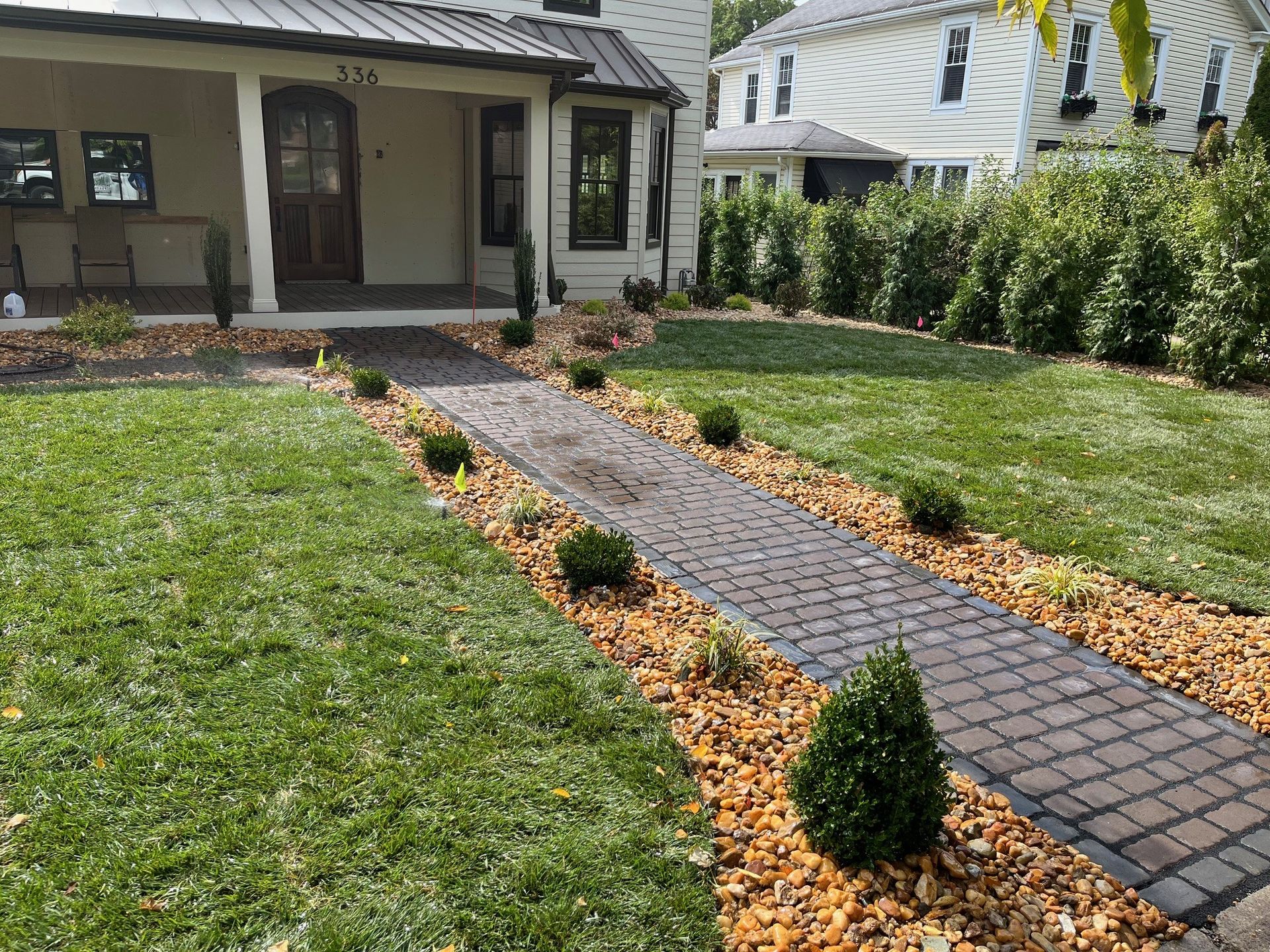 A brick walkway leads to a covered front porch of a white house, flanked by mulch beds with small evergreen shrubs.