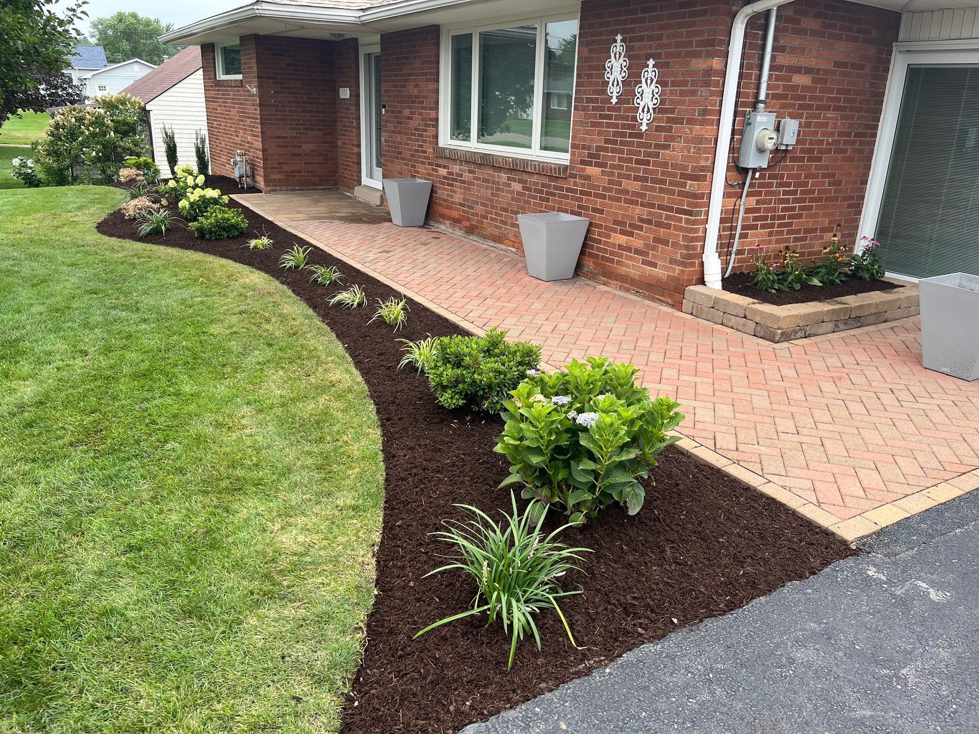 A brick ranch house features a front yard with a curved mulch garden bed containing green shrubs beside a paved walkway.