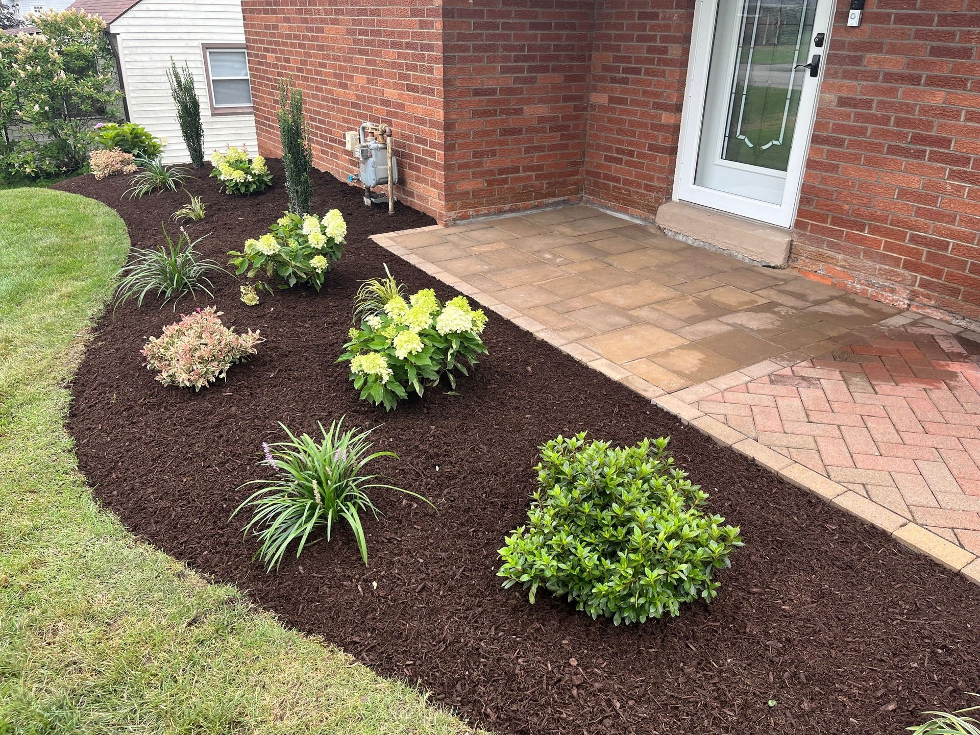 A brick house entrance with a stone porch surrounded by a mulched garden bed featuring various small green plants.