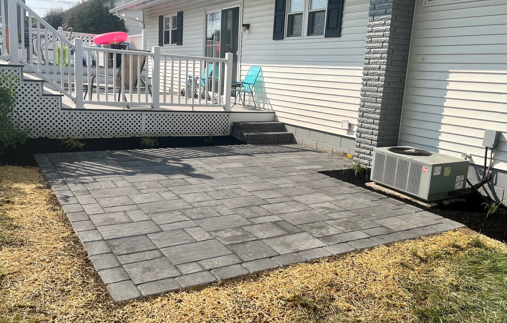 A new gray paver patio set on the ground next to a white house with a raised wooden deck and an HVAC unit.