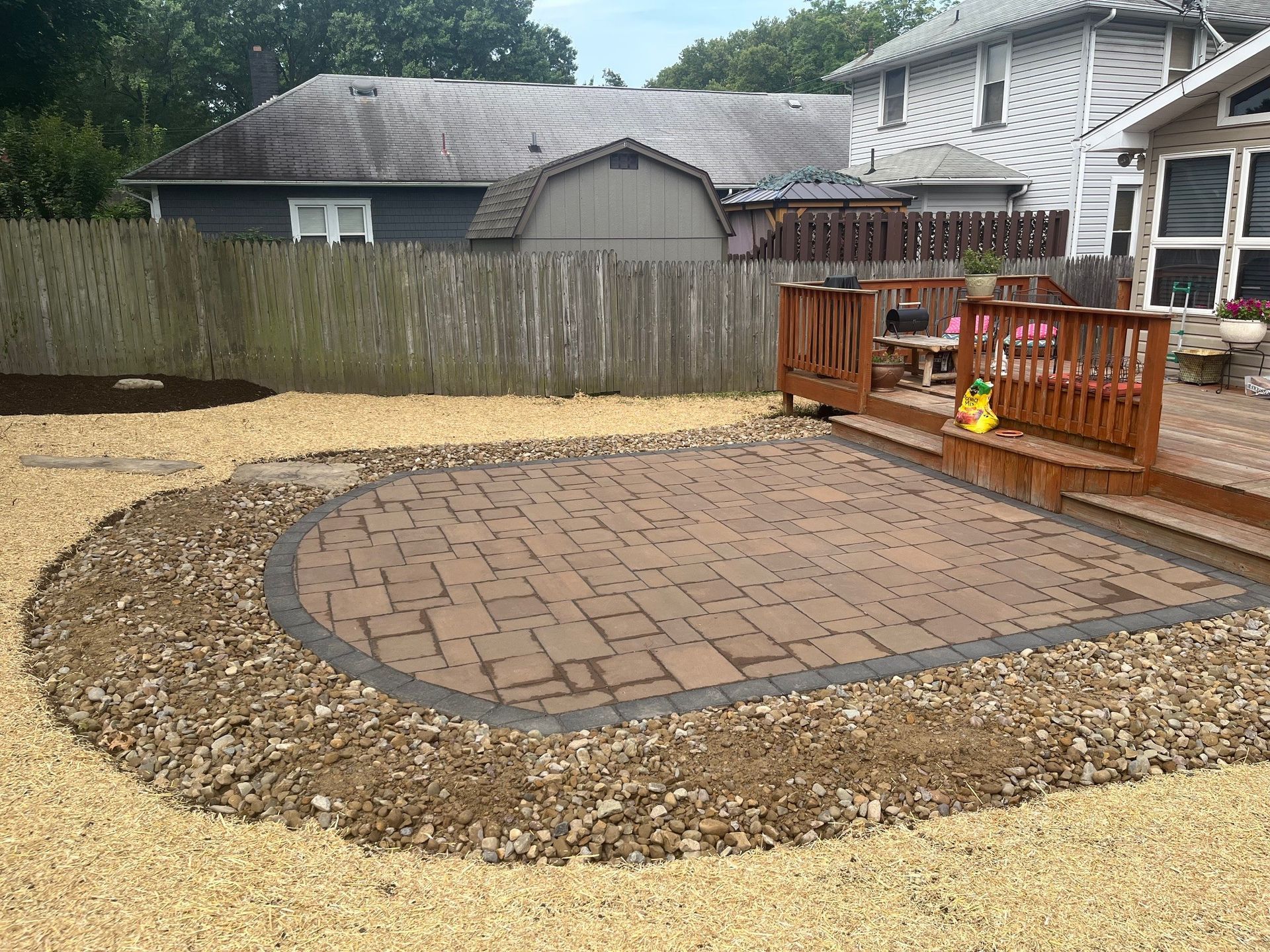 A backyard with a circular brick patio, surrounded by gravel, next to a wooden deck and fence.