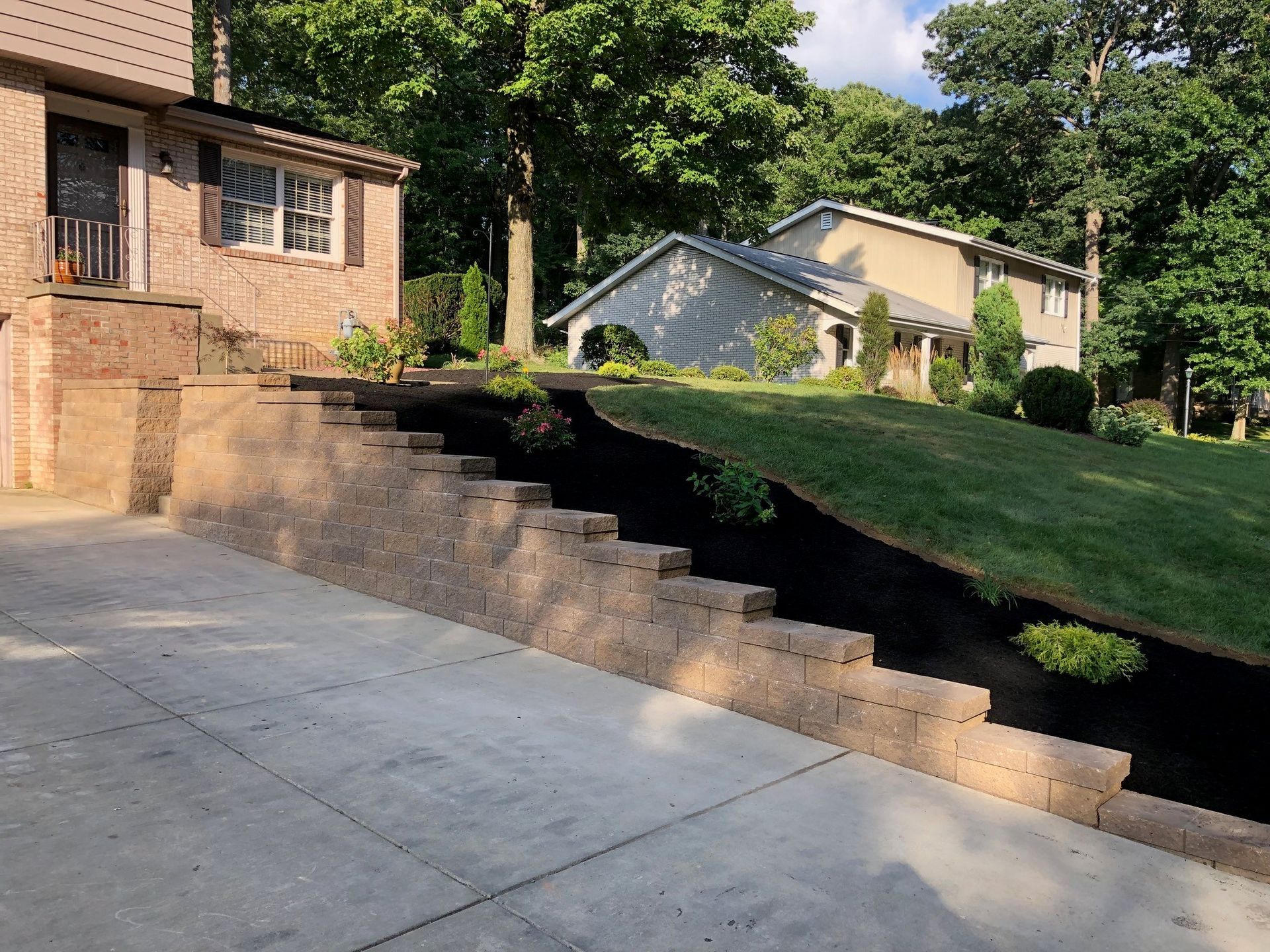 A tan, tiered retaining wall runs alongside a concrete driveway, holding up a sloped green lawn in a suburban yard.